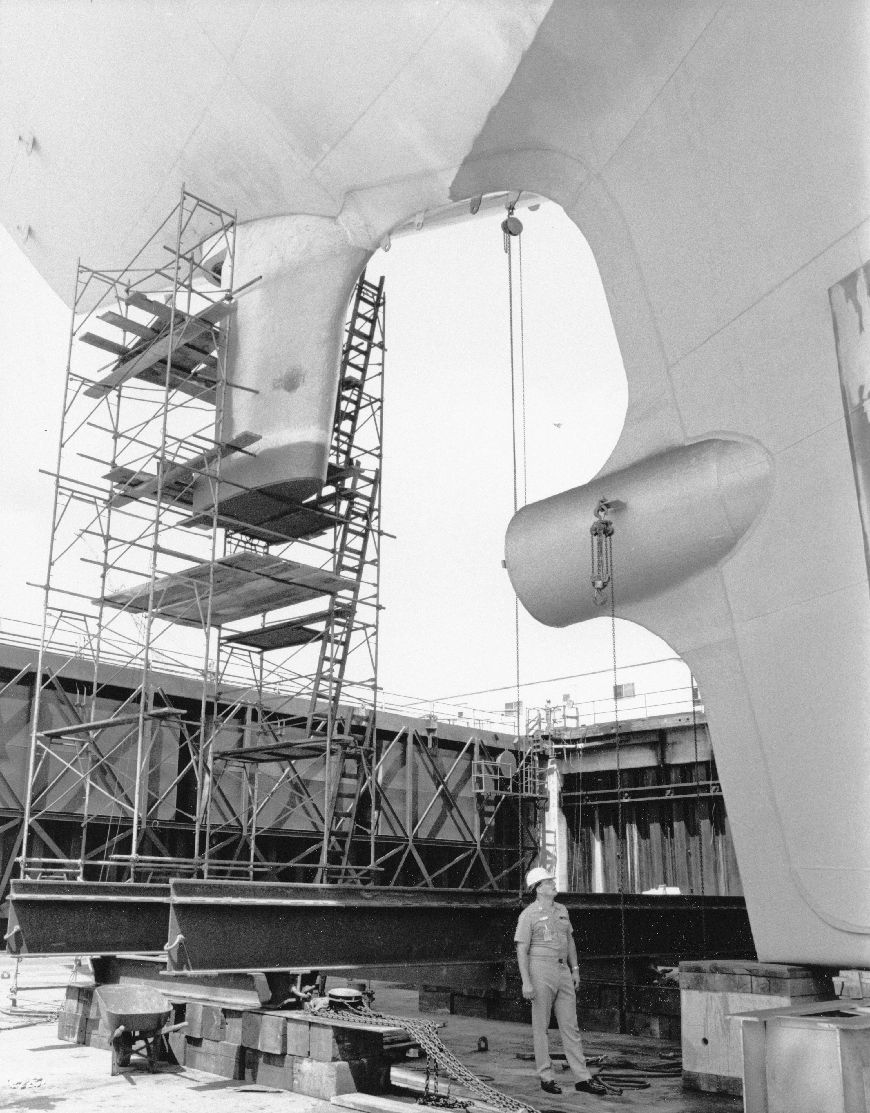 A shipyard worker beneath a large ship propeller and rudder.